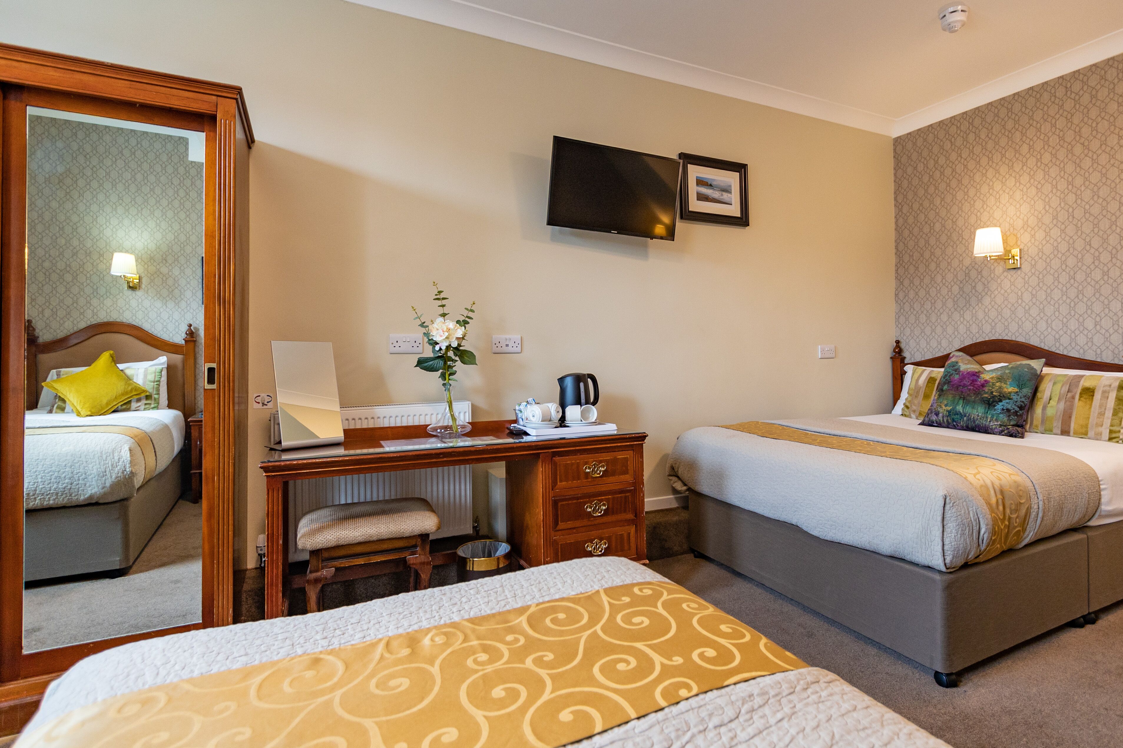 A hotel room with two beds, a desk with a chair, a mirror, a television, and a small table with a flower vase.