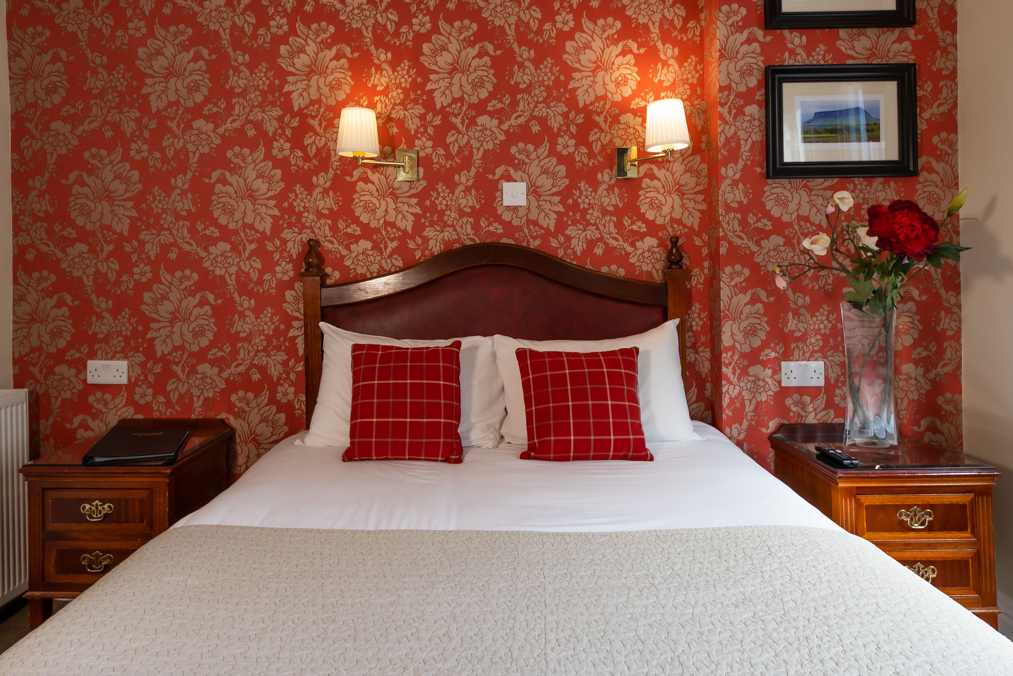 Une chambre d’hôtel avec un grand lit, un papier peint à motifs rouges et blancs, deux lampes fixées au mur, une table de chevet avec un téléphone et un vase de fleurs placé sur la table d’appoint.