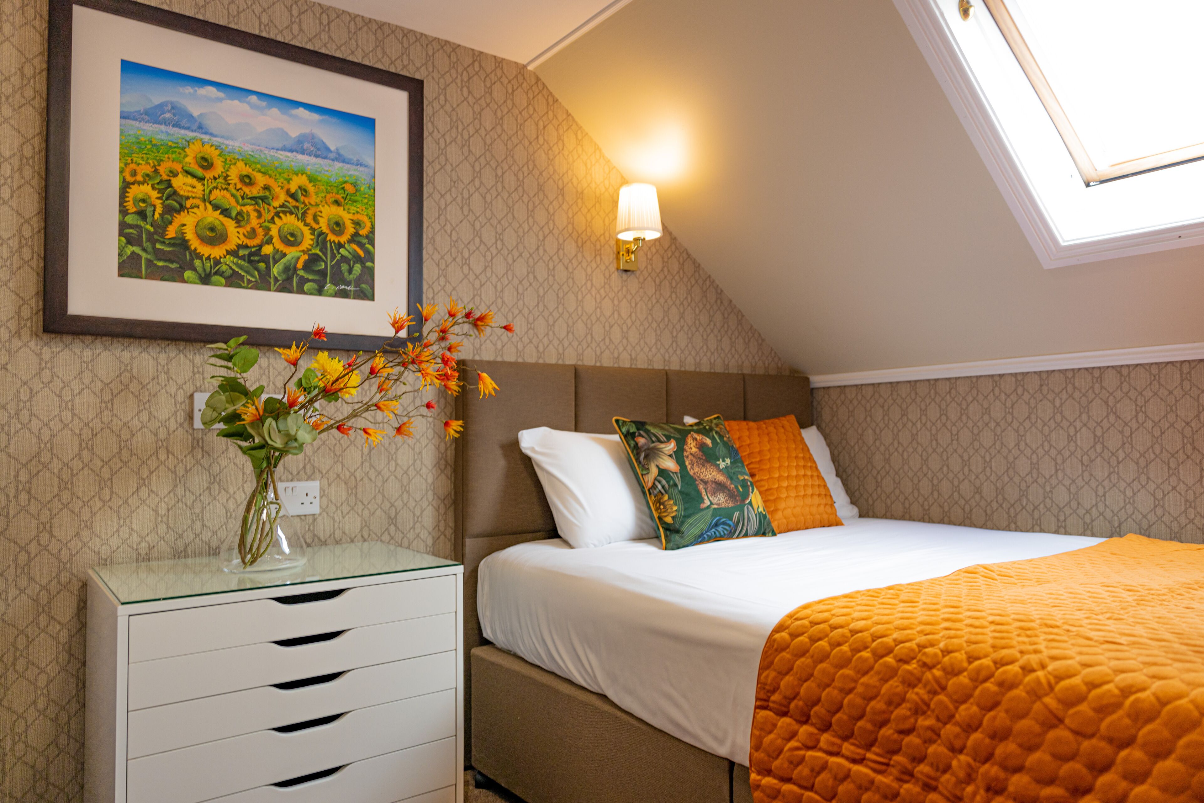 A bedroom with a bed, a nightstand, a vase with flowers, a wall-mounted lamp, and a framed painting of sunflowers.