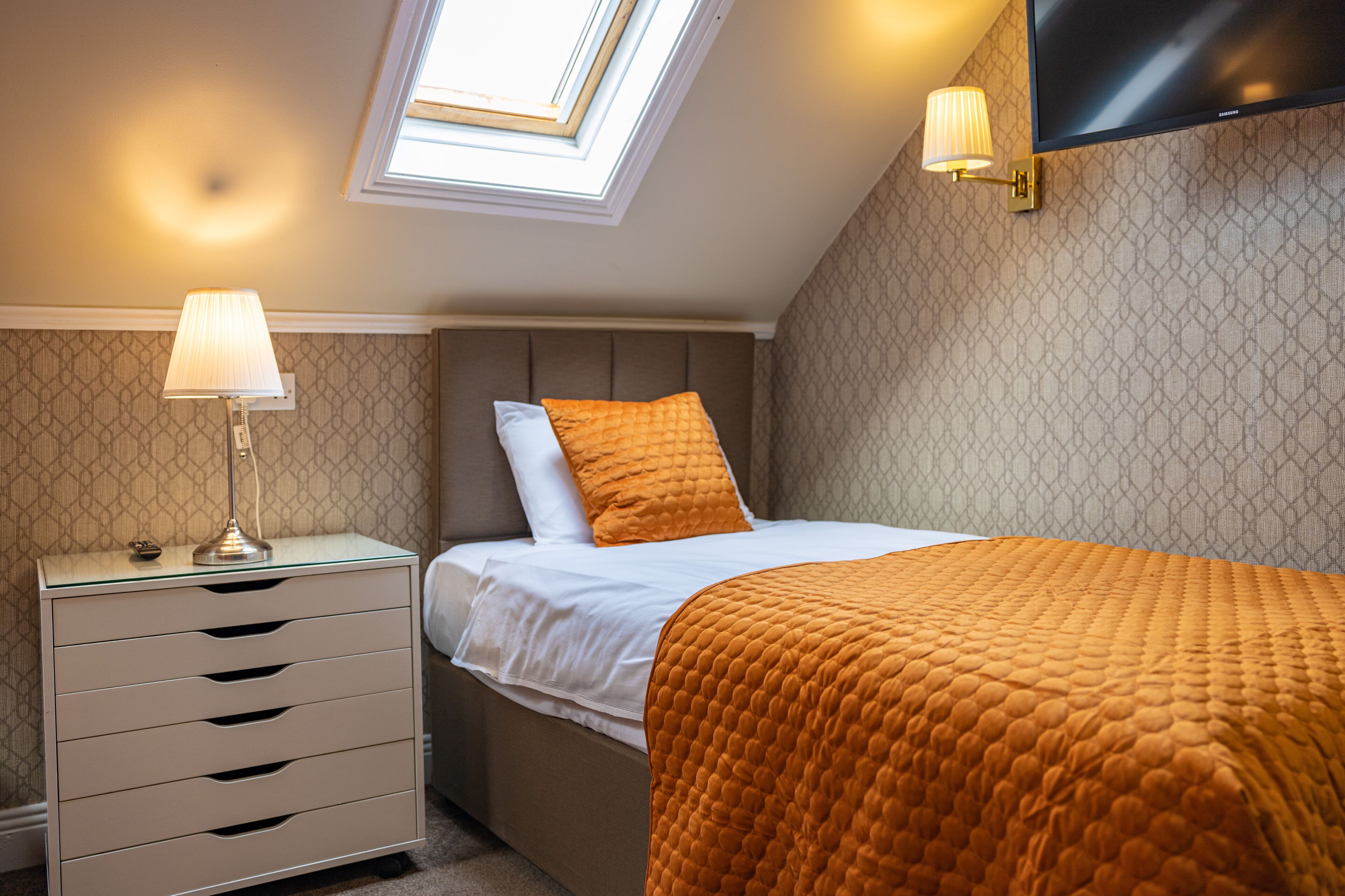 Ein Schlafzimmer mit einem Bett, einem Nachttisch, einer Lampe, einem Fernseher und einem Dachfenster.