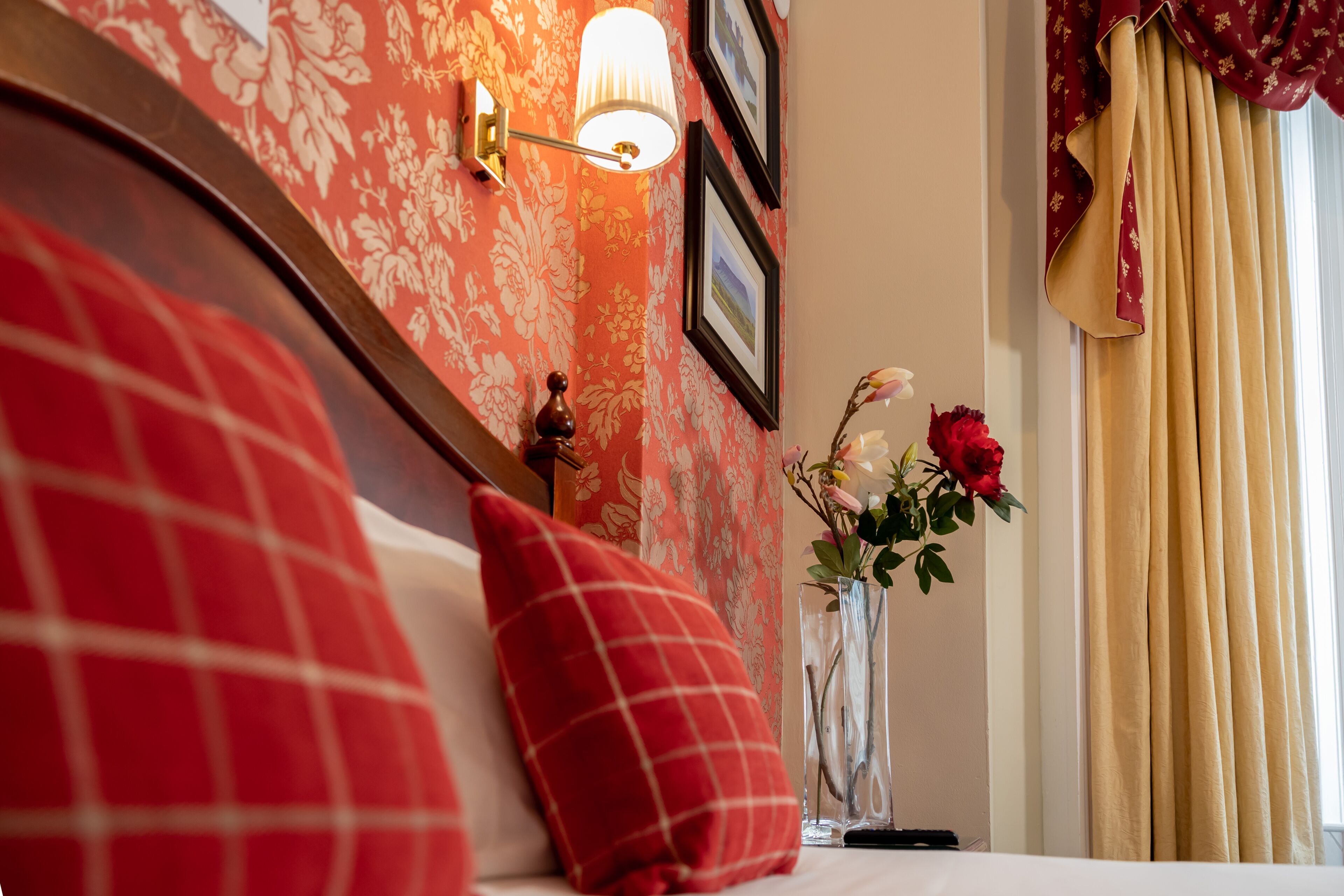 A hotel room with floral wallpaper, a bed with red and white pillows, a bedside table with a vase of flowers, and a window with curtains.