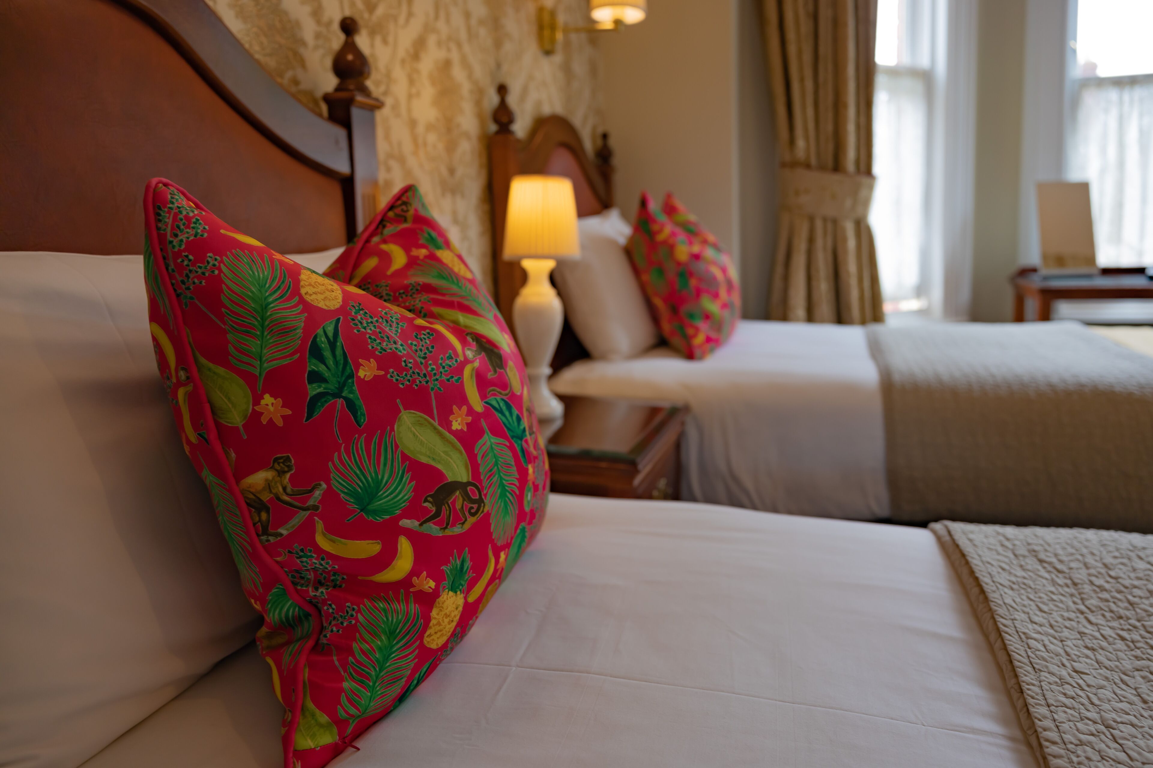 A hotel room with two beds, each with white linens and colorful tropical-themed pillows. There is a wooden headboard, a bedside table with a lamp, and a window with curtains.