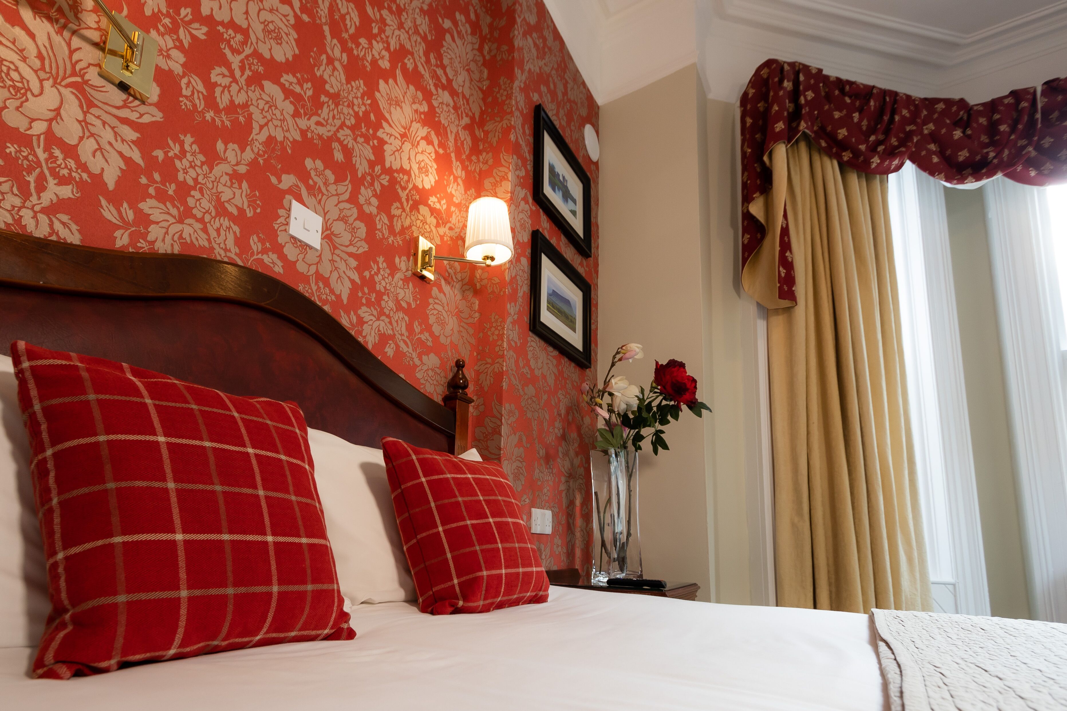 A neatly made bed with red and white checkered pillows, a vase with flowers, and a wall with floral wallpaper and framed pictures.