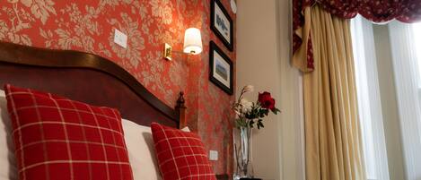 A neatly made bed with red and white checkered pillows, a vase with flowers, and a wall with floral wallpaper and framed pictures.