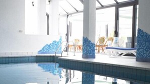 Una piscina interior con paredes y techo azulejados, paneles de vidrio transparente y zonas iluminadas por el sol.