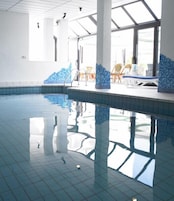 Una piscina interior con paredes y techo azulejados, paneles de vidrio transparente y zonas iluminadas por el sol.