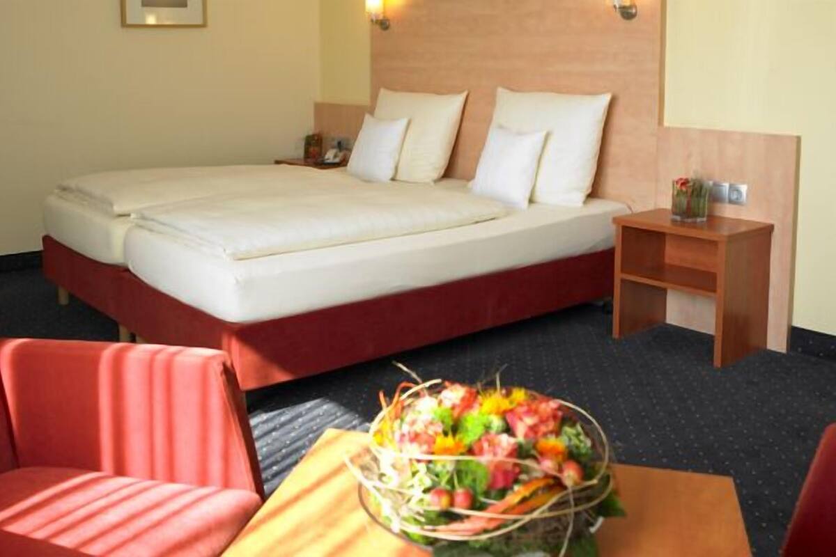 A hotel room with a bed, two red chairs, a wooden table, and a fruit basket.