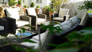 Un área de estar al aire libre con mobiliario de mimbre y cojines blancos, rodeada de plantas en macetas y una mesa de vidrio.