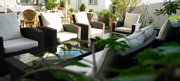 Un área de estar al aire libre con mobiliario de mimbre y cojines blancos, rodeada de plantas en macetas y una mesa de vidrio.