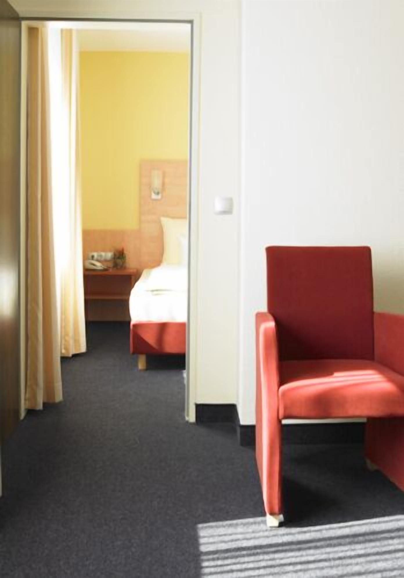 Ein Hotelzimmer mit einem roten Sessel, einem Bett mit weißer Bettwäsche und einem Nachttisch aus Holz mit einer Lampe.