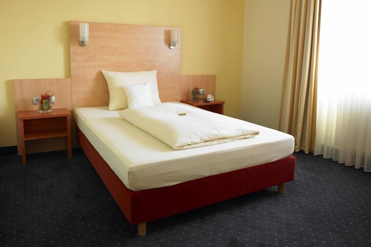 A hotel room with a single bed, wooden headboard, two bedside tables with lamps, and a window with curtains.
