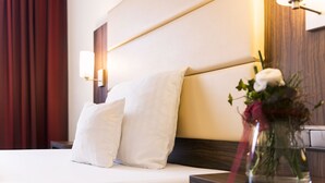 Una cama bien hecha con sábanas y almohadas blancas, una mesita de noche con un florero y una lámpara de pared.