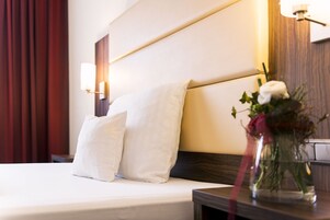 Una cama bien hecha con sábanas y almohadas blancas, una mesita de noche con un florero y una lámpara de pared.