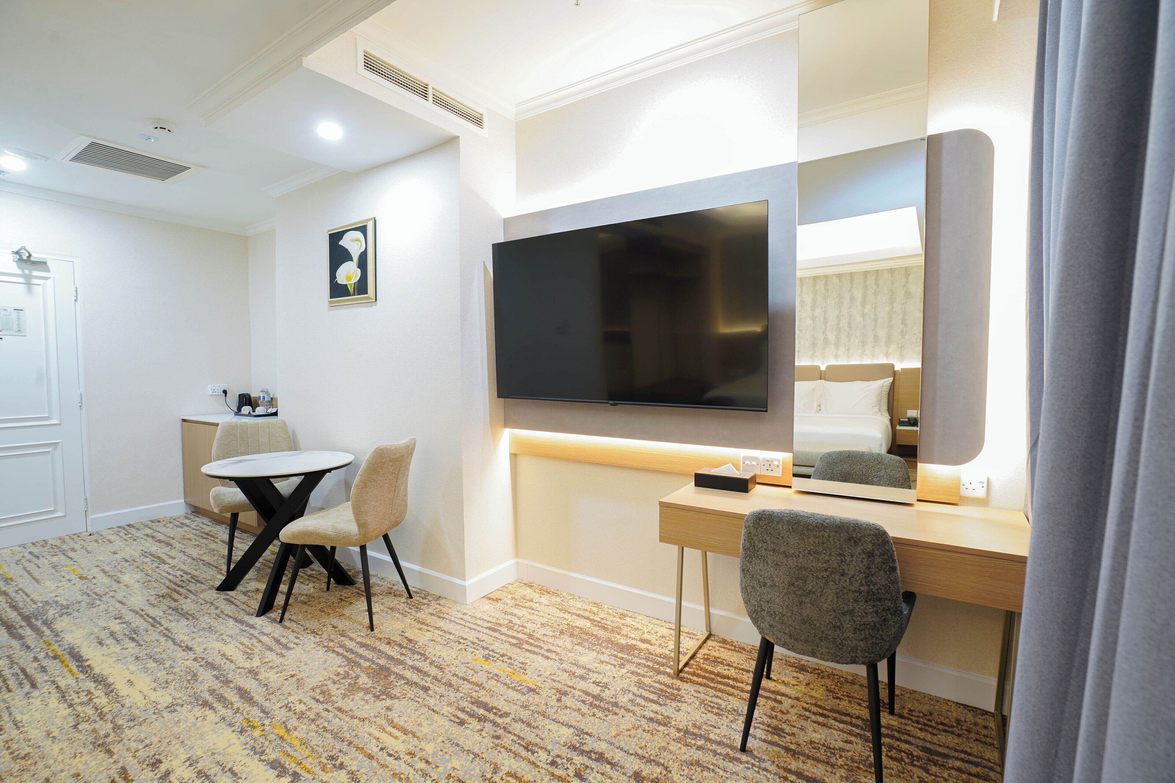 A hotel room with a flat-screen TV, a desk, two chairs, a small table, and a carpeted floor.