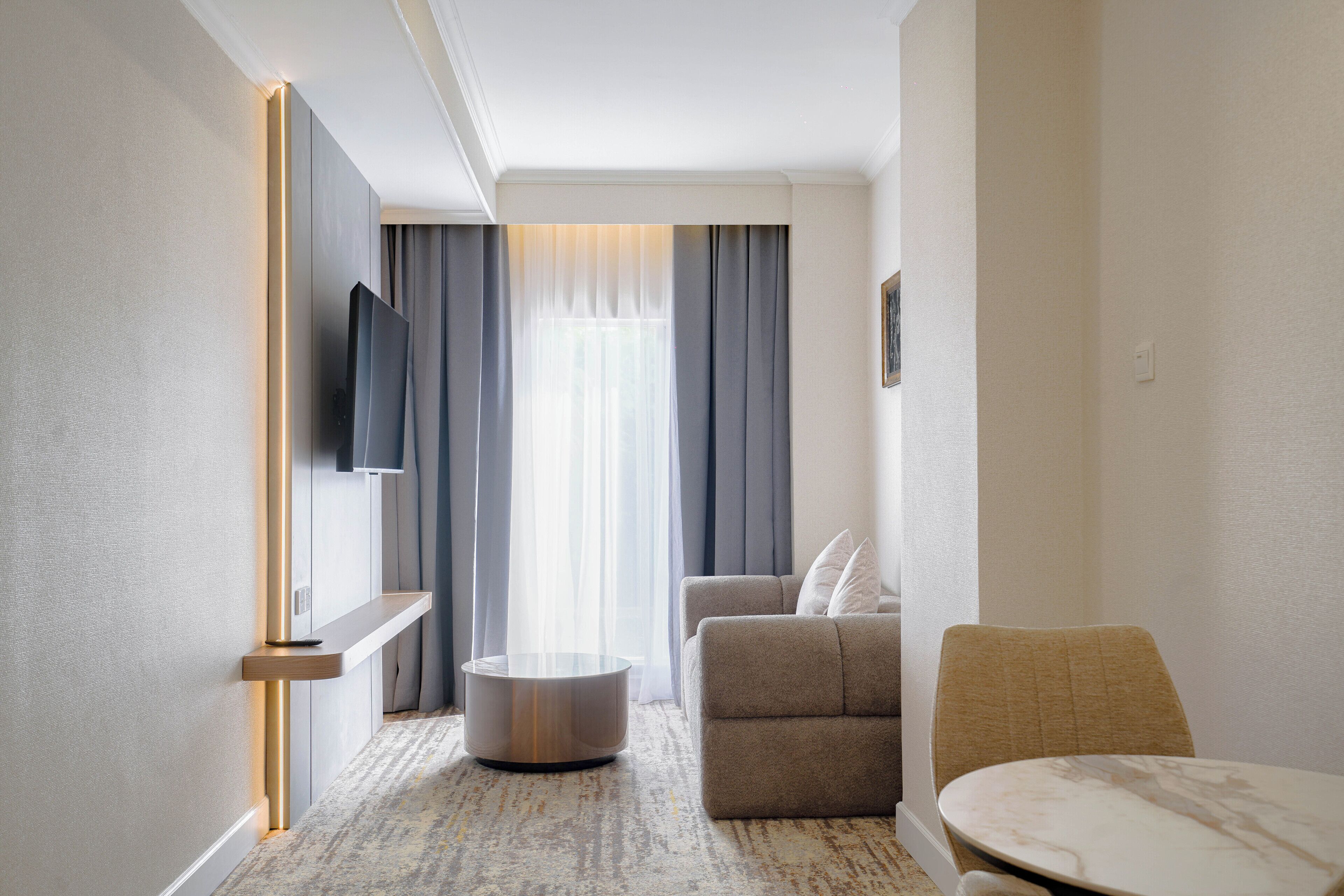 A hotel room with a flat-screen TV mounted on the wall, a round coffee table, a sofa with cushions, and a small round table with a chair.
