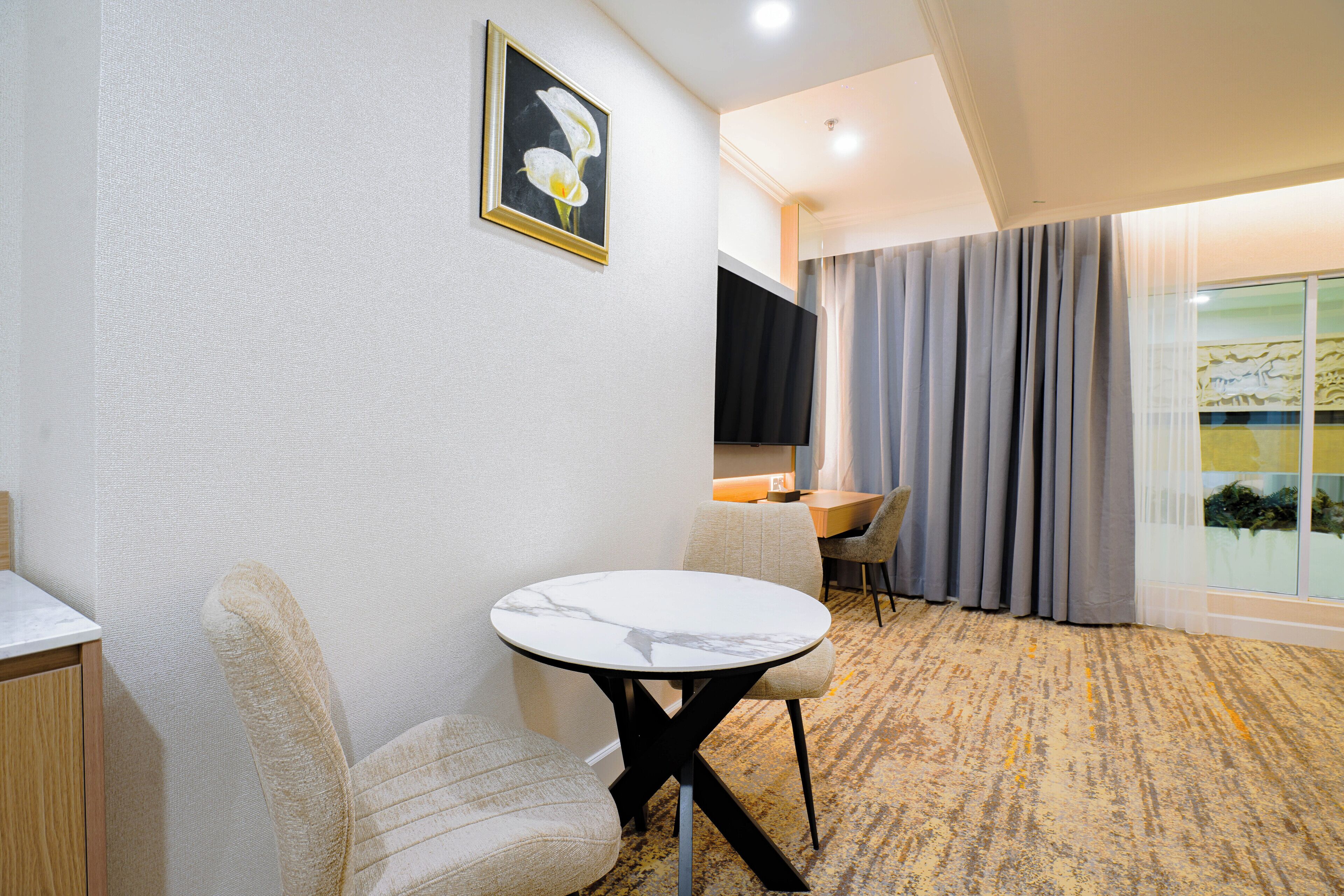 Ein Hotelzimmer mit rundem Tisch, einer Couch, einem Fernseher und einem großen Fenster mit Vorhängen.