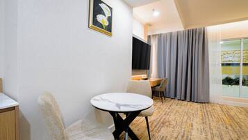 Ein Hotelzimmer mit rundem Tisch, einer Couch, einem Fernseher und einem großen Fenster mit Vorhängen.
