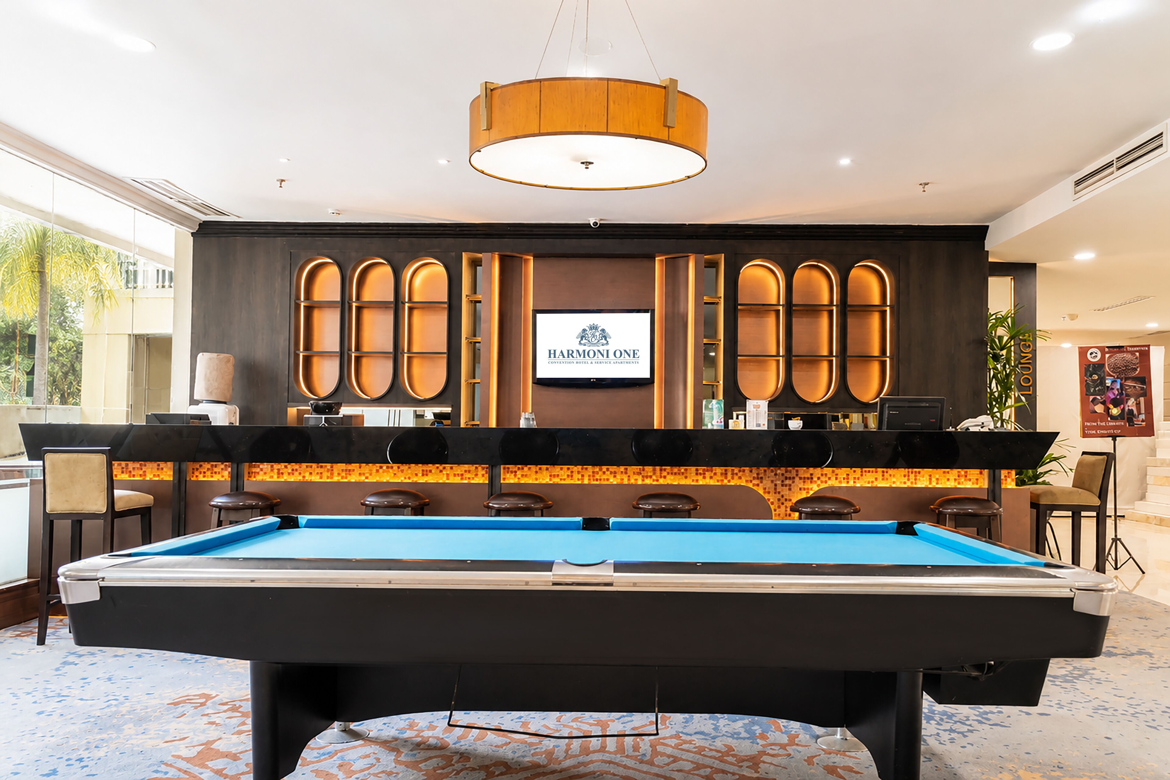A pool table with a billiards cue rack, a television screen displaying 'HARMONI ONE', and a bar area with stools.