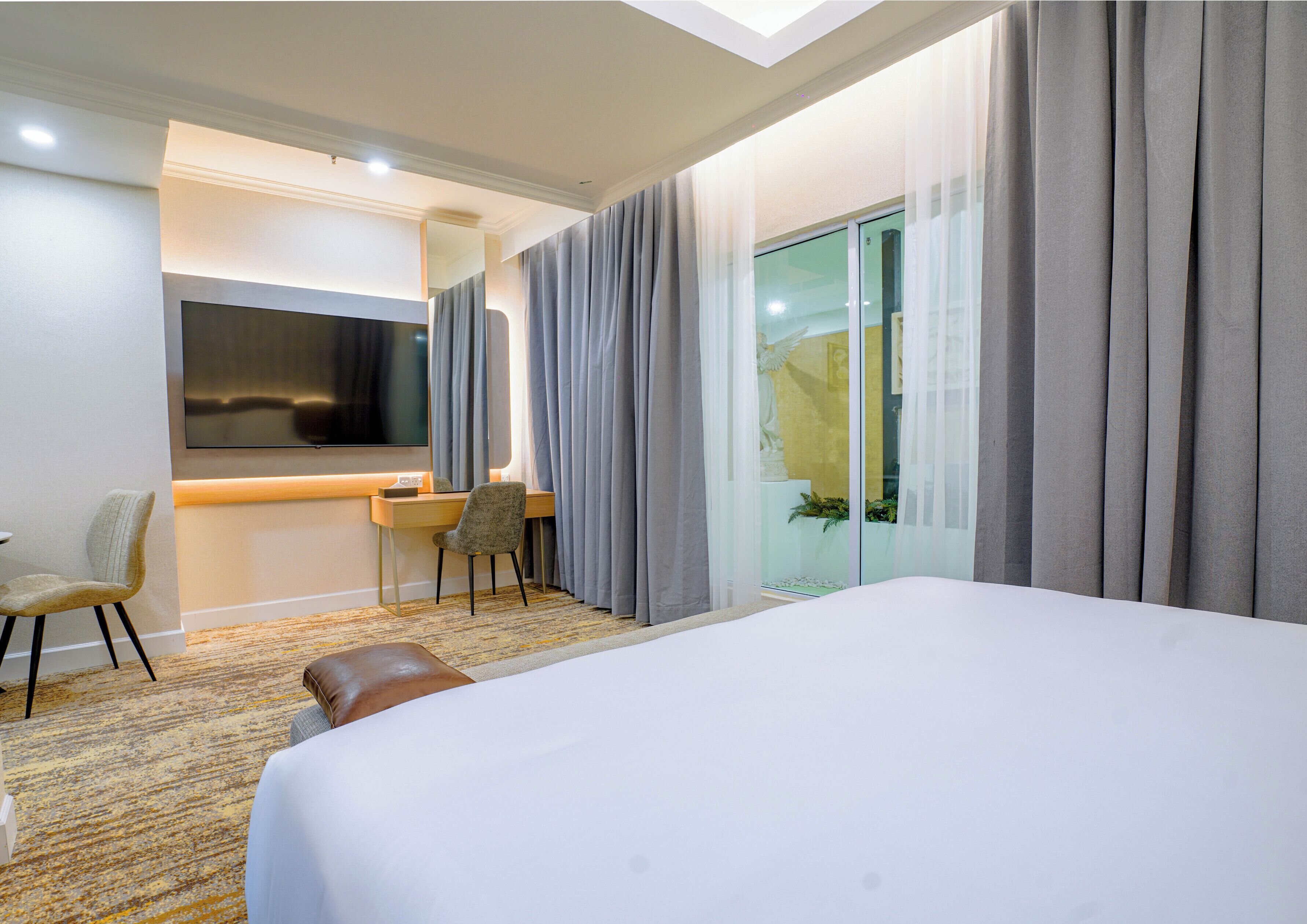 Habitación de hotel con cama, escritorio con silla, televisor y ventana grande con cortinas.