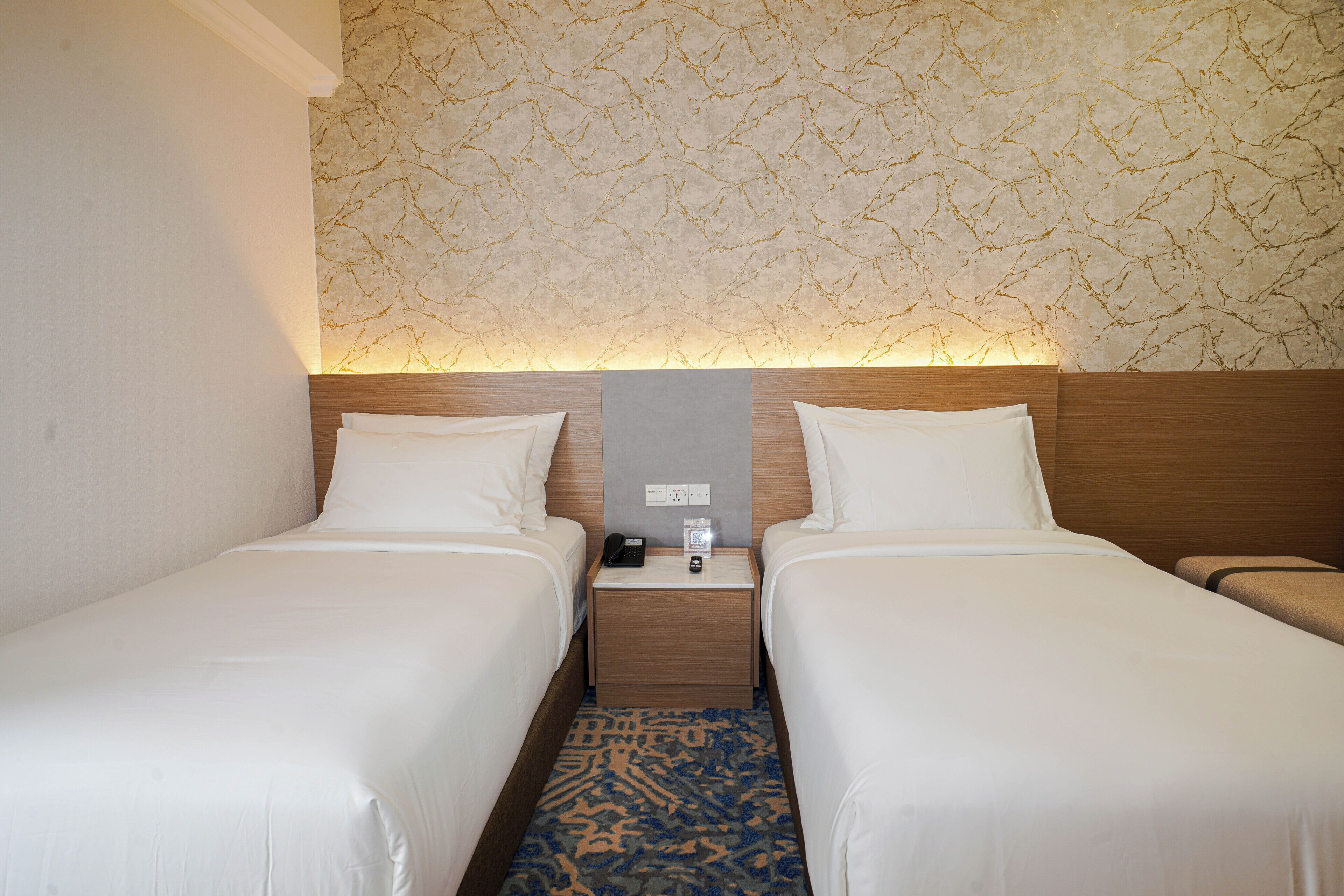 A hotel room with two single beds, a nightstand with a phone and a remote, and a patterned wall.