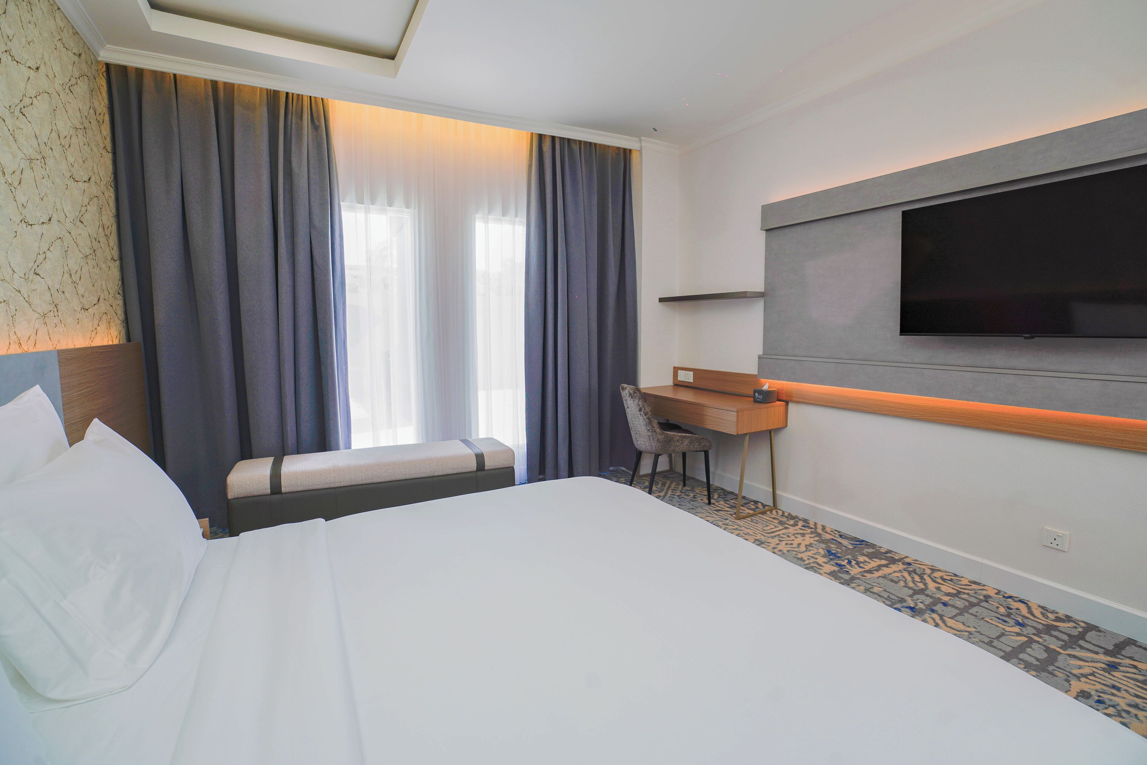 Habitación de hotel con cama, escritorio, silla, televisor y ventana con cortinas.