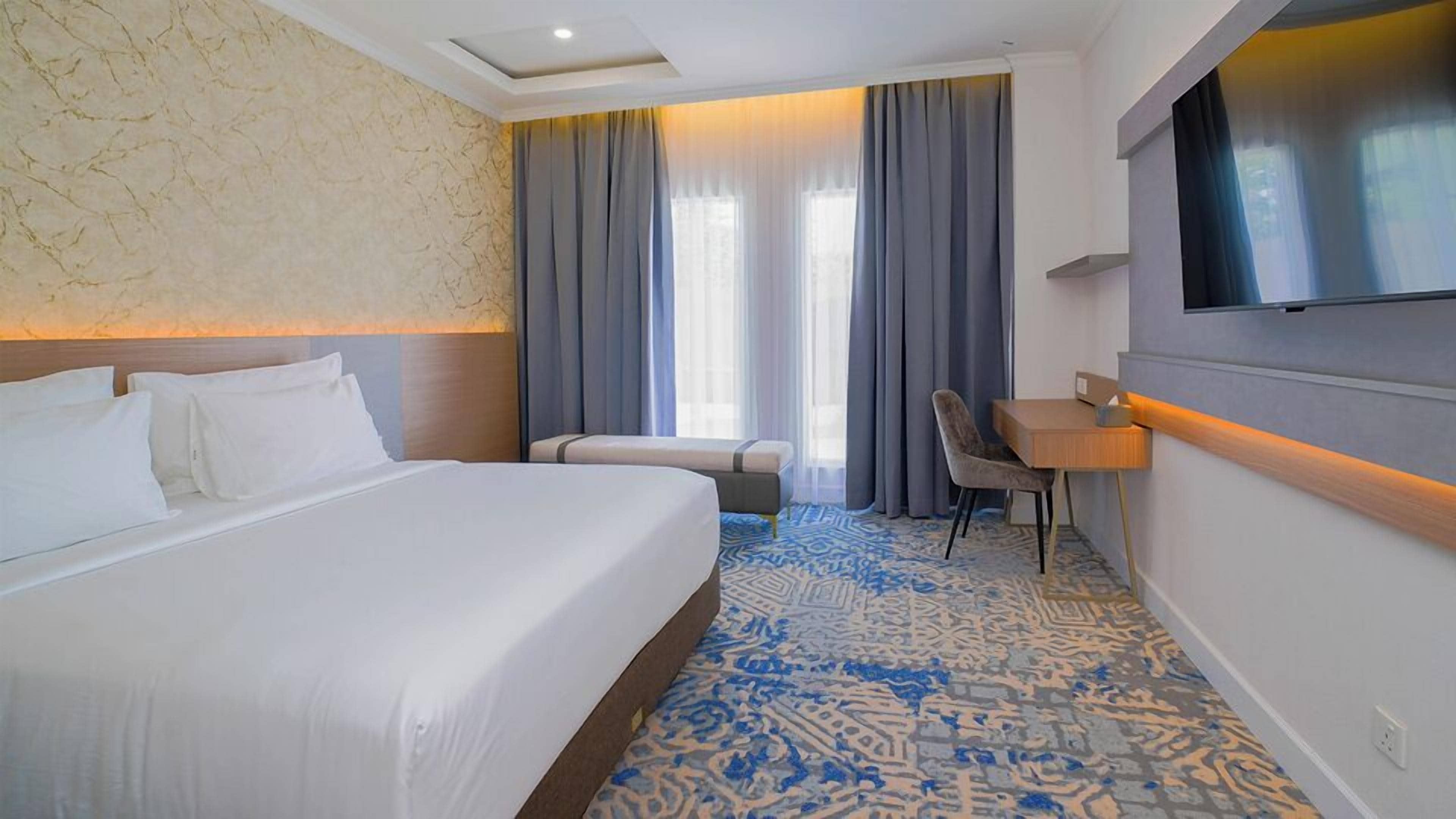 Habitació d'hotel amb un llit gran, una taula de treball, una cadira, televisió i una finestra amb cortines.