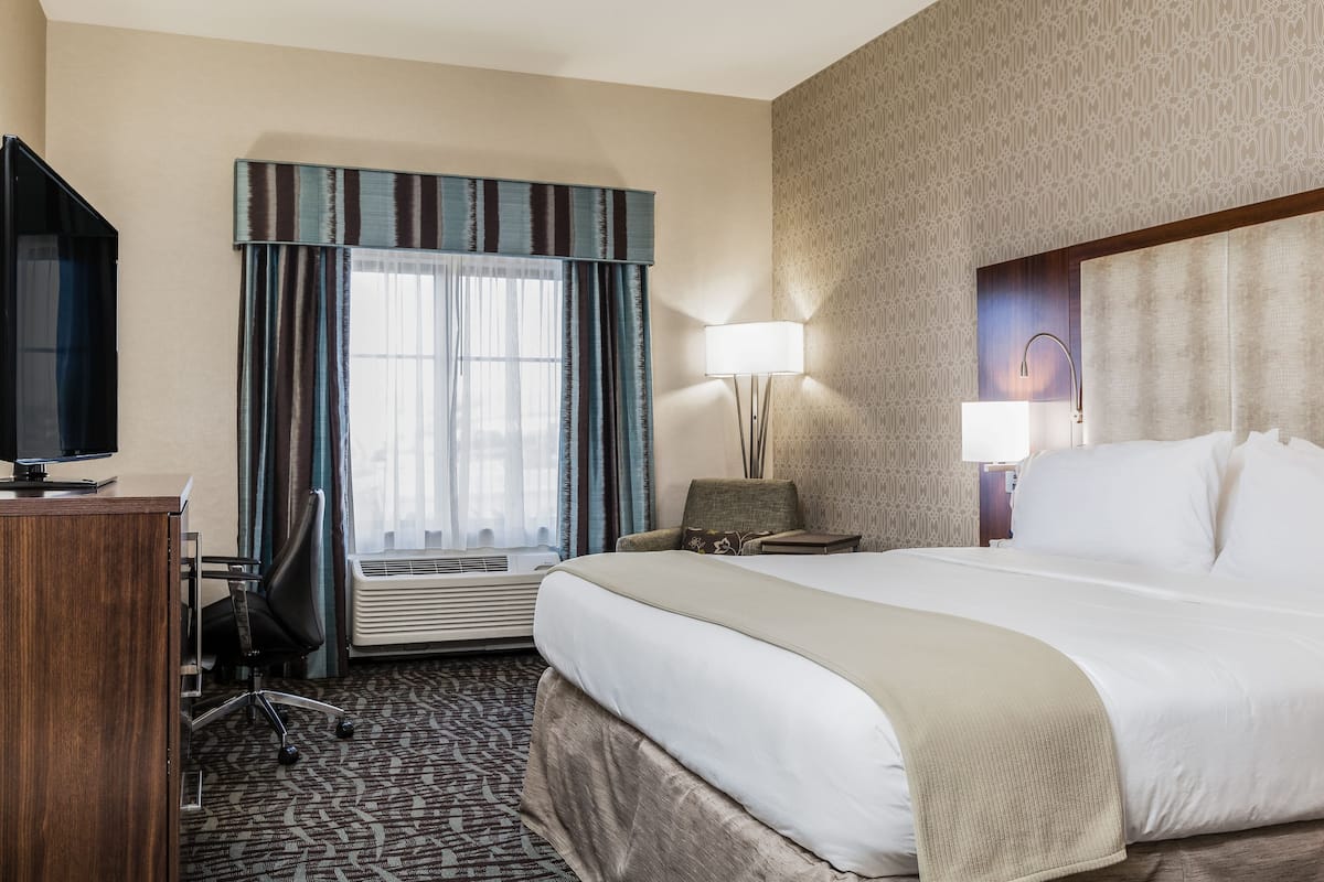 A hotel room with a large bed, a desk, a chair, a TV, a lamp, and a window with curtains.