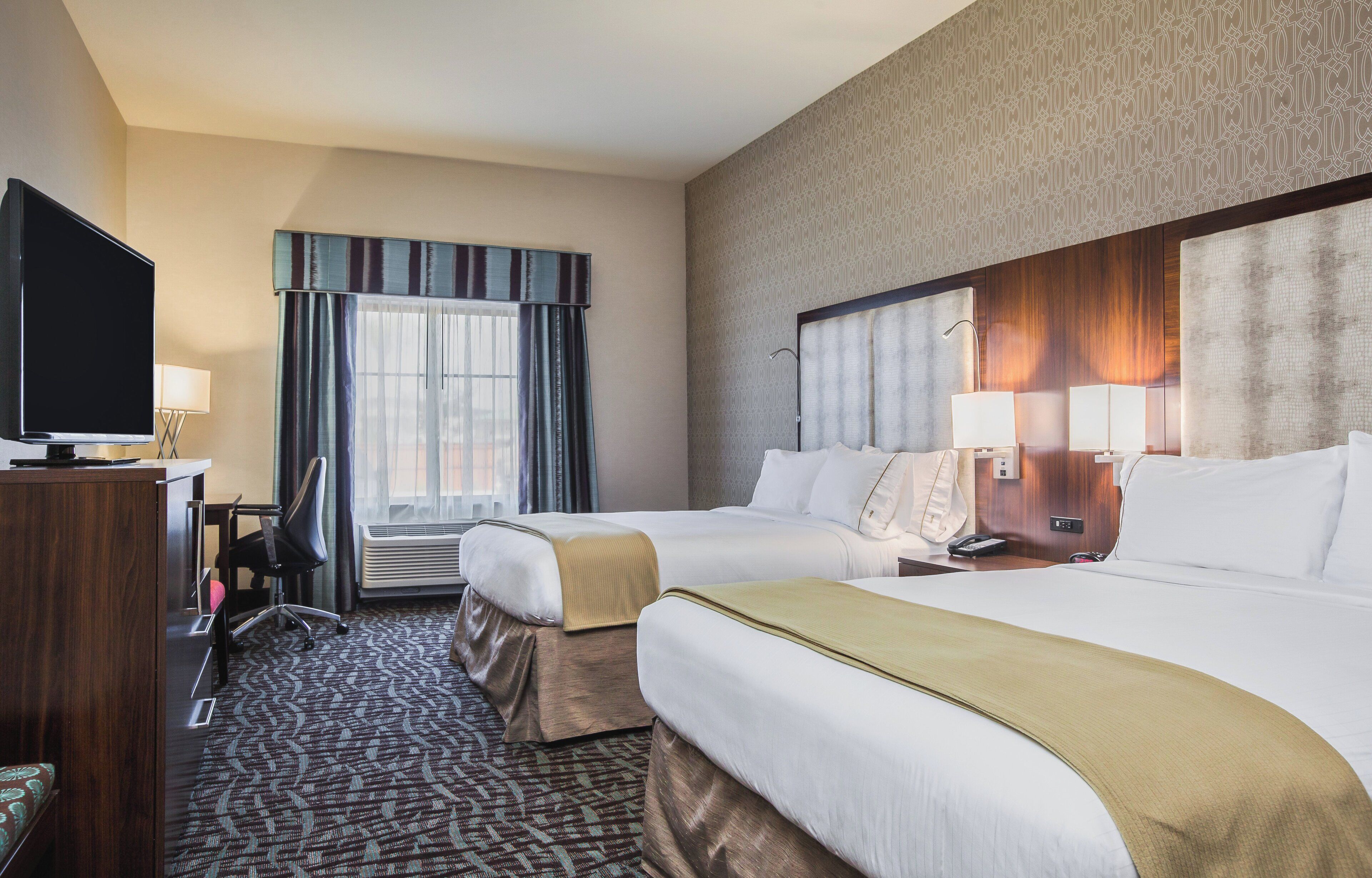 Habitación de hotel con dos camas, un televisor, un escritorio con lámpara y una alfombra estampada.