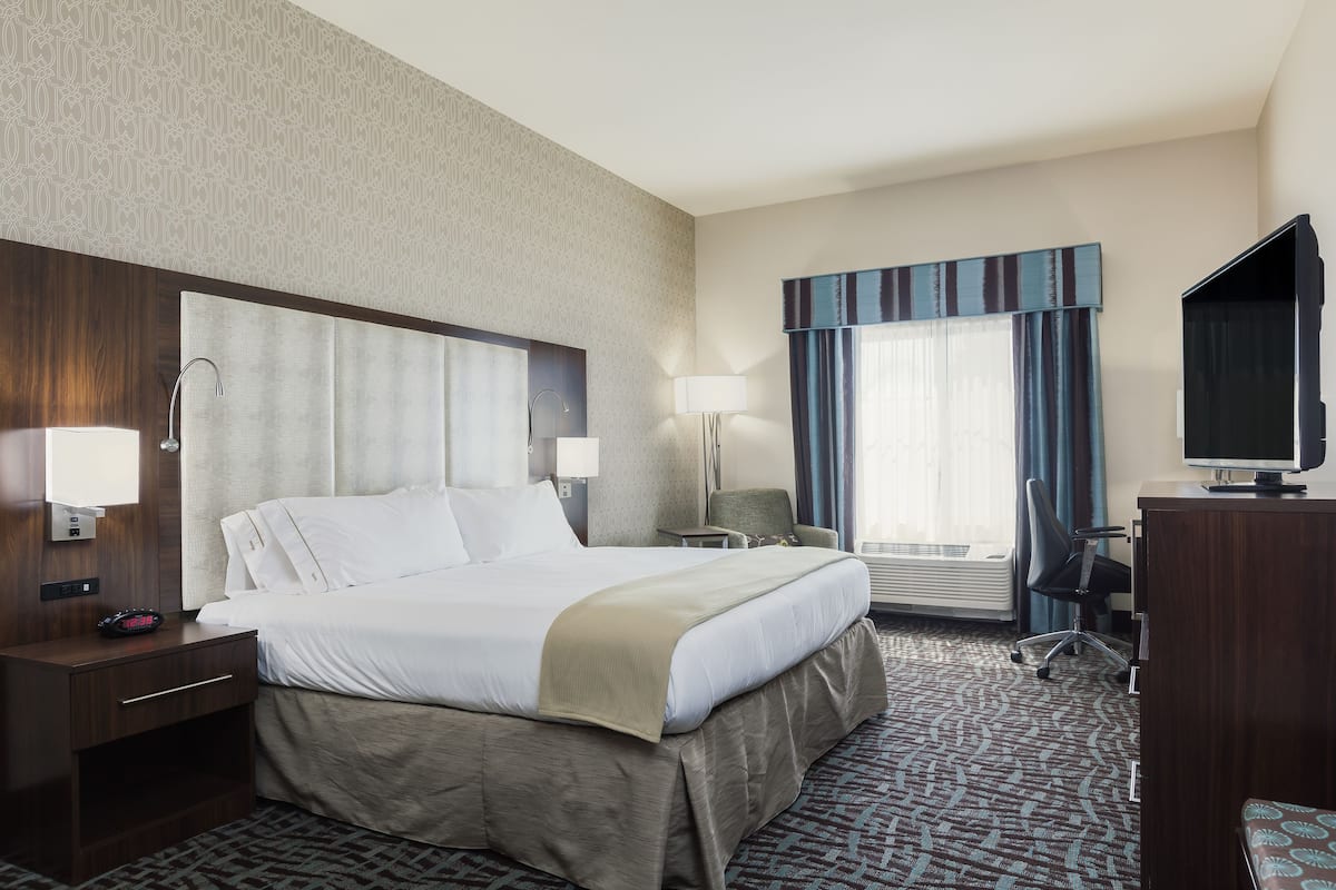 A hotel room with a large bed, bedside tables, a desk, a chair, a television, and a window with curtains.