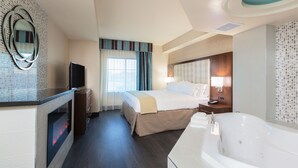 A hotel room with a large bed, a TV, a fireplace, a bathroom with a bathtub, and a mirror on the wall.