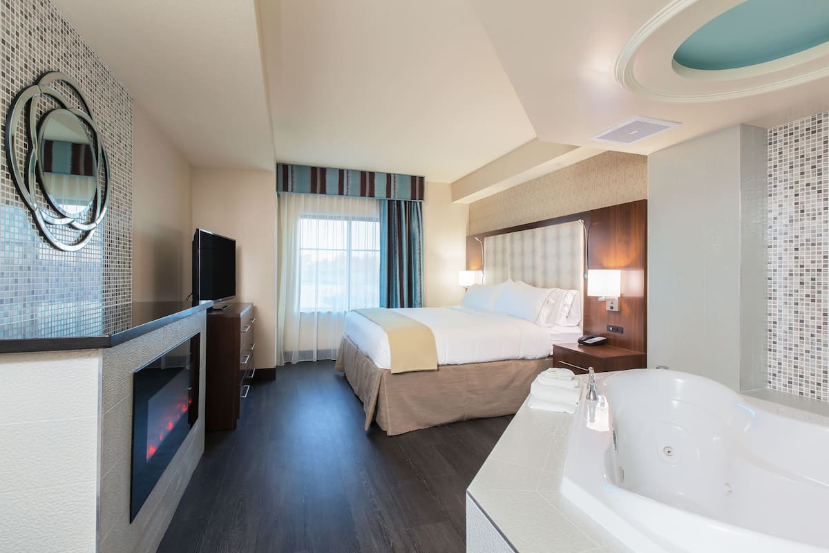 A hotel room with a large bed, a TV, a fireplace, a bathroom with a bathtub, and a mirror on the wall.