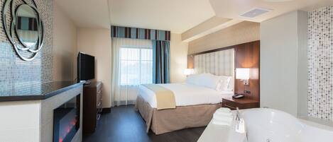 A hotel room with a large bed, a TV, a fireplace, a bathroom with a bathtub, and a mirror on the wall.