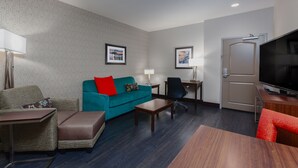 A hotel room with a sofa, armchair, ottoman, coffee table, TV, and a door.