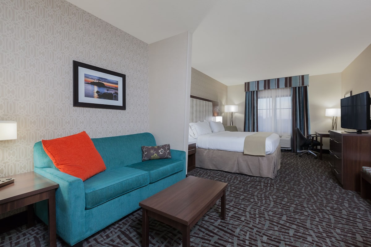 A hotel room with a teal sofa, a wooden coffee table, a bed with a white comforter, a desk with a television, and a patterned carpet.