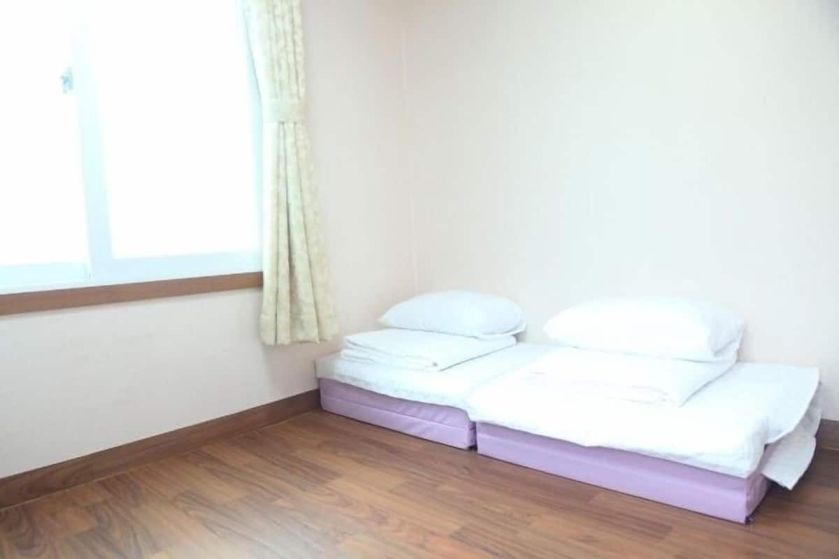 Una cama pequeña y bien hecha con ropa de cama blanca en una habitación con piso de madera y una ventana con cortinas.