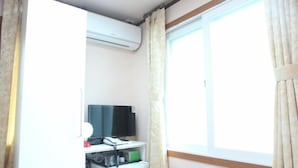 Una habitación con ventana, aire acondicionado, escritorio con computadora y teléfono, y un refrigerador pequeño.