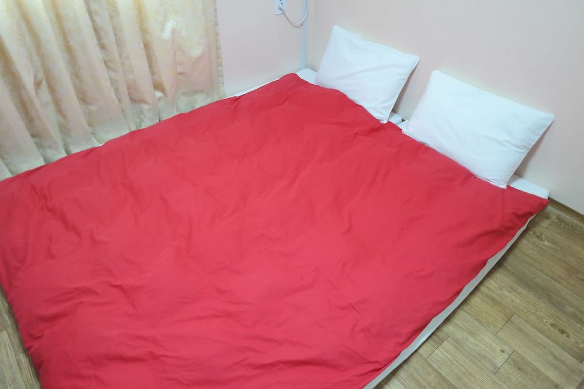 Una cama con una colcha roja y almohadas blancas en una habitación con suelo de madera y pared de color claro.