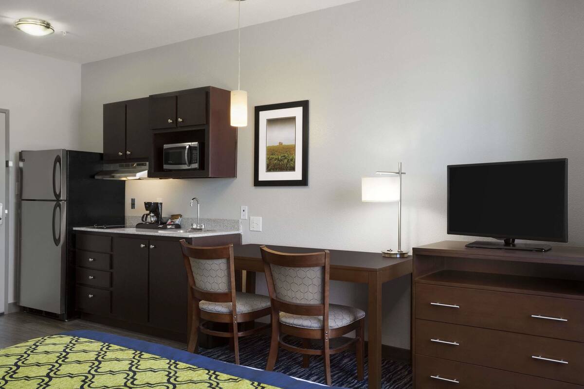 Een compacte hotelkamer met een kitchenette, een eethoek met twee stoelen, een flatscreen televisie en een ingelijst kunstwerk aan de muur.