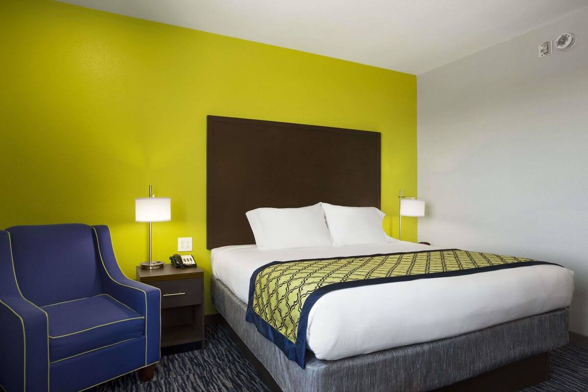 Een hotelkamer met een bed, een blauwe fauteuil, een nachtkastje met een lamp, en een heldergele accentmuur.