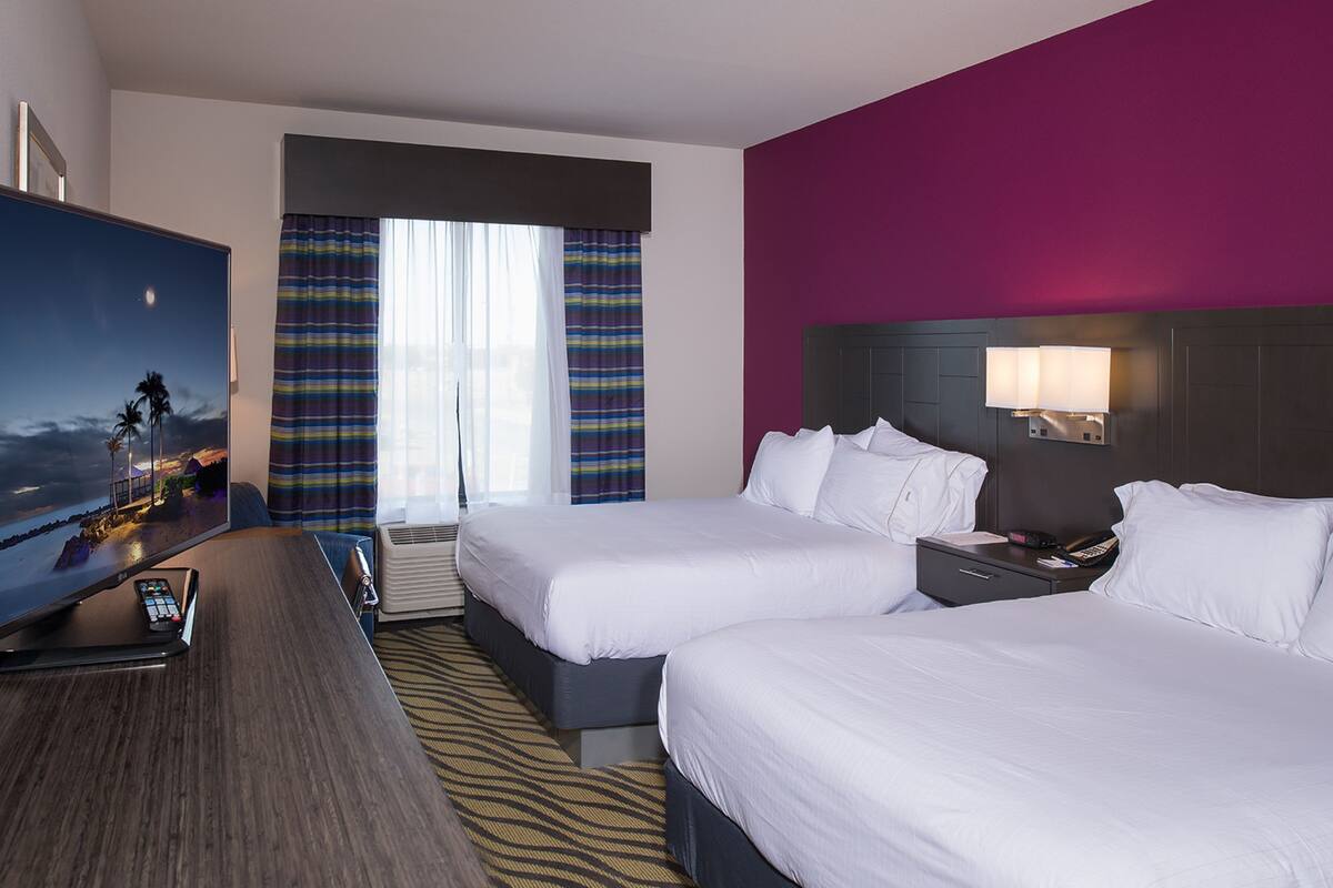 A hotel room with two beds, a flat-screen TV, a desk, and a window with curtains.