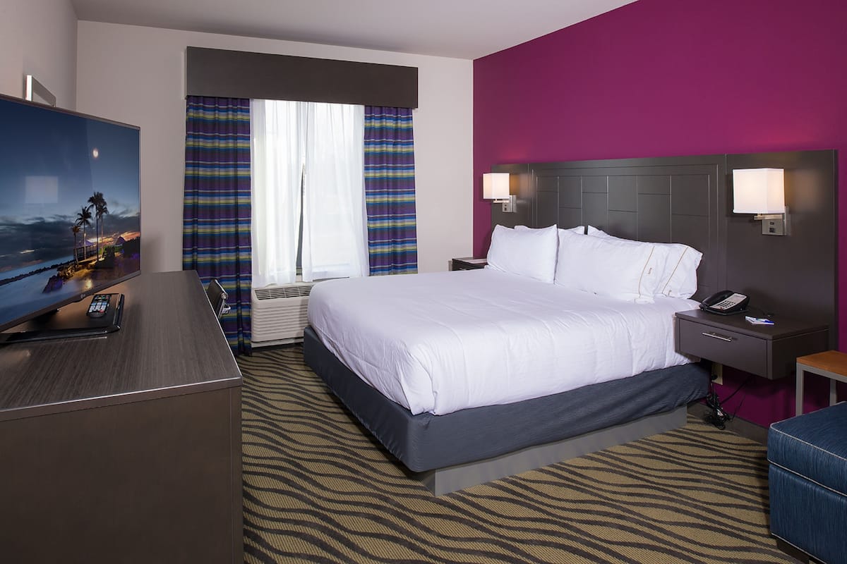 A hotel room with a large bed, a desk with a flat-screen TV, a nightstand with a phone, and a window with curtains.