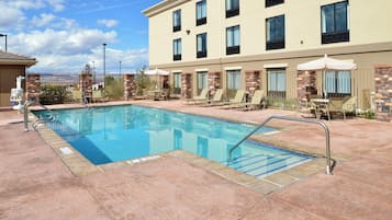 Una piscina con sillas de descanso y sombrillas, rodeada por un edificio con múltiples ventanales y detalles de piedra.