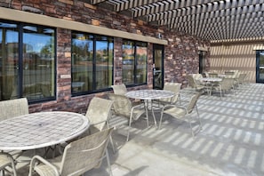 An outdoor patio area with round tables and wicker chairs, a brick wall, and large windows. - Holiday Inn Express & Suites Page - Lake Powell Area by IHG (Page)