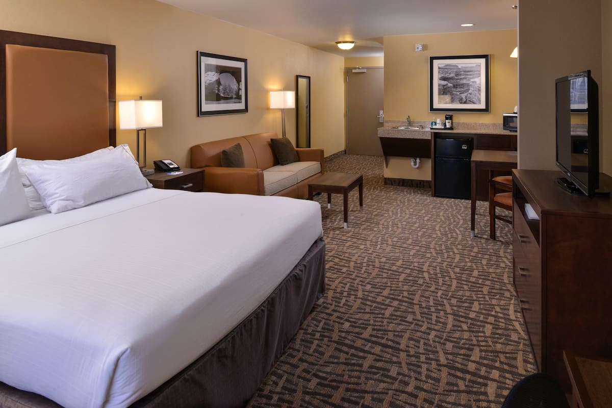 A hotel room with a large bed, a sofa, a desk, a television, and a small kitchenette.