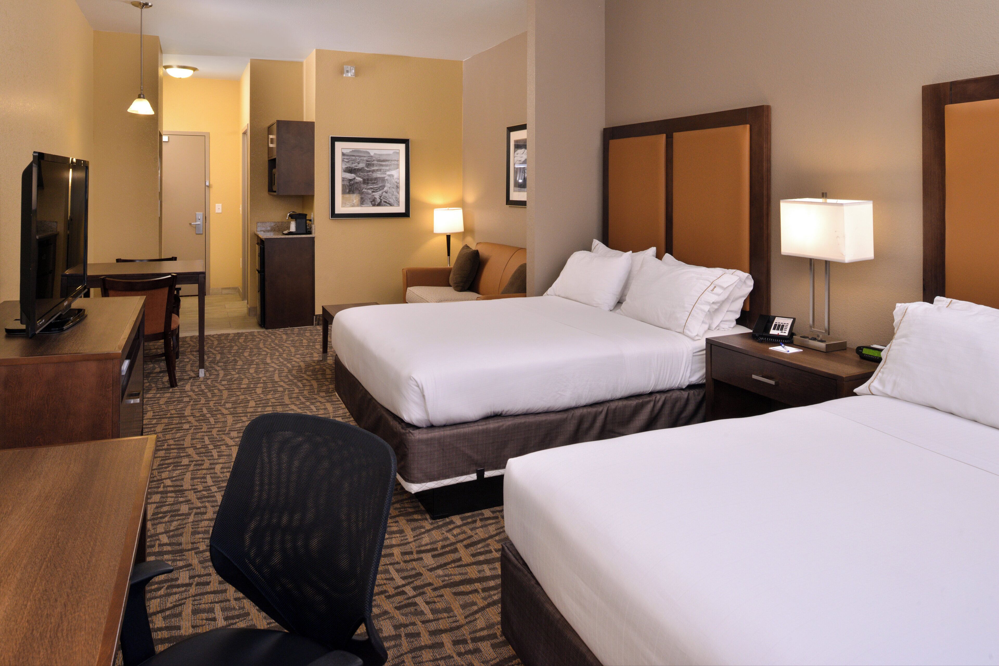 A hotel room with two beds, a desk with a chair, a television, a lamp, and framed pictures on the wall.