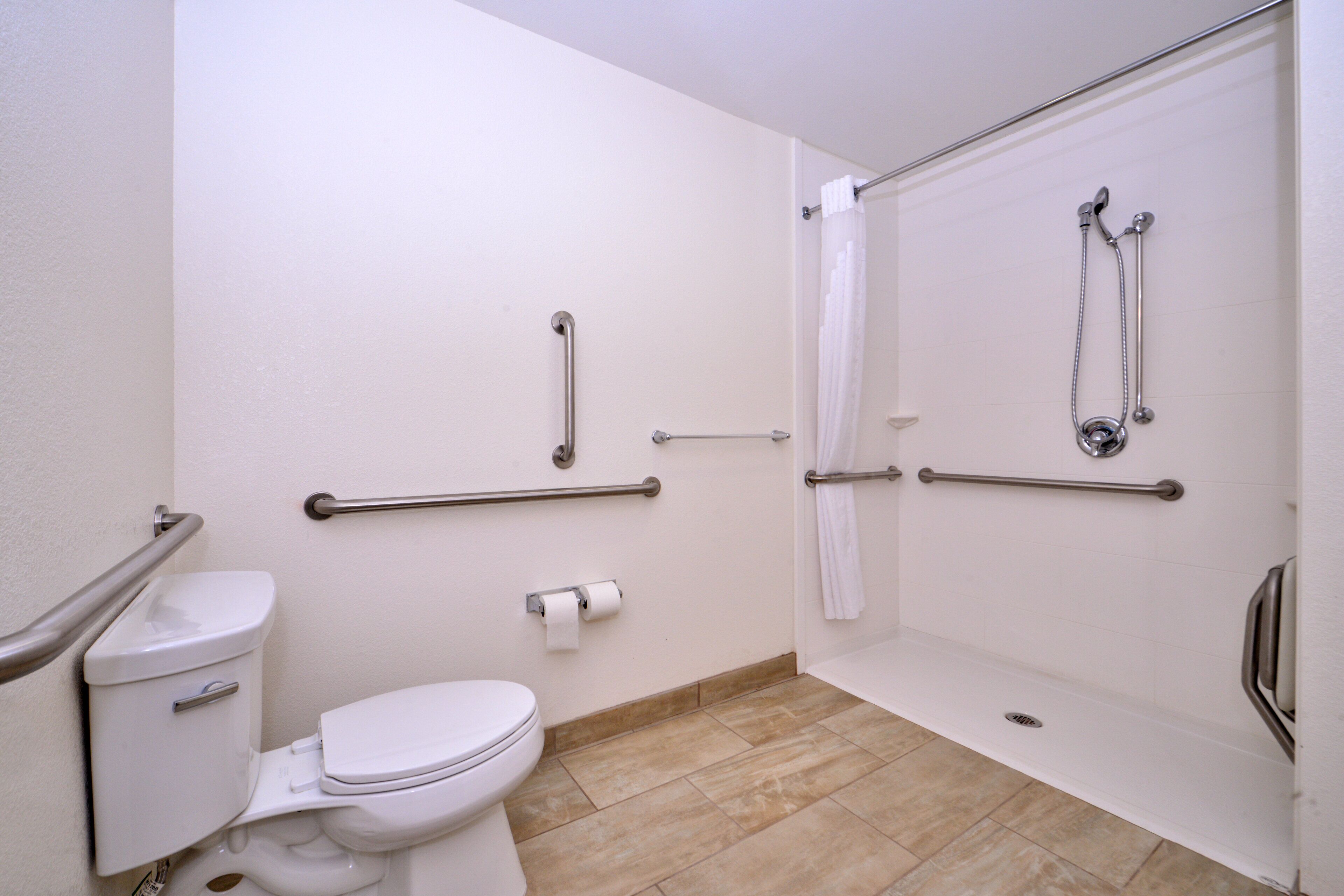 Une salle de bain équipée de toilettes, d’une douche avec des barres d’appui et d’un rouleau de papier toilette fixé au mur.