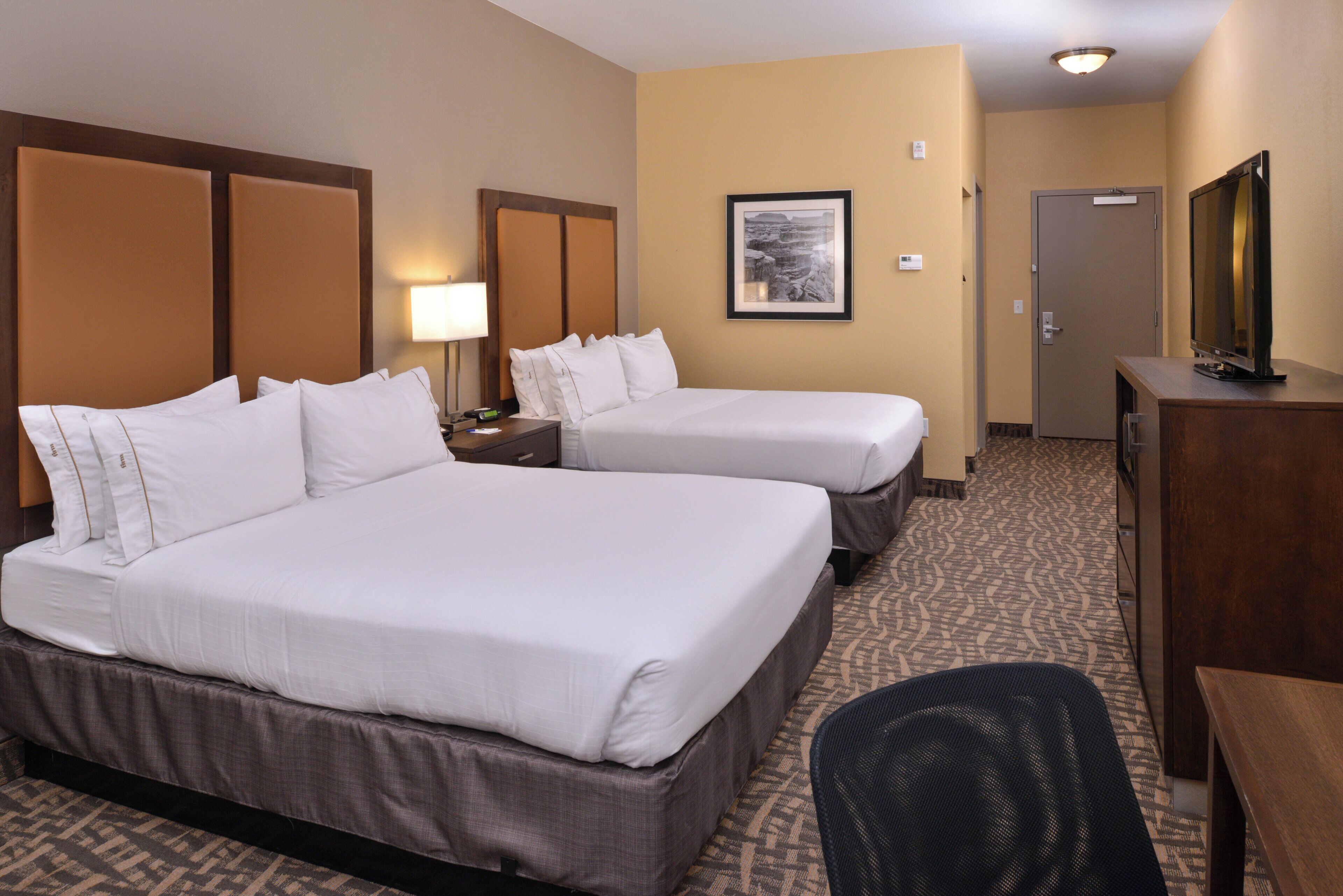 A hotel room with two beds, a desk, a chair, a lamp, and a framed picture on the wall.