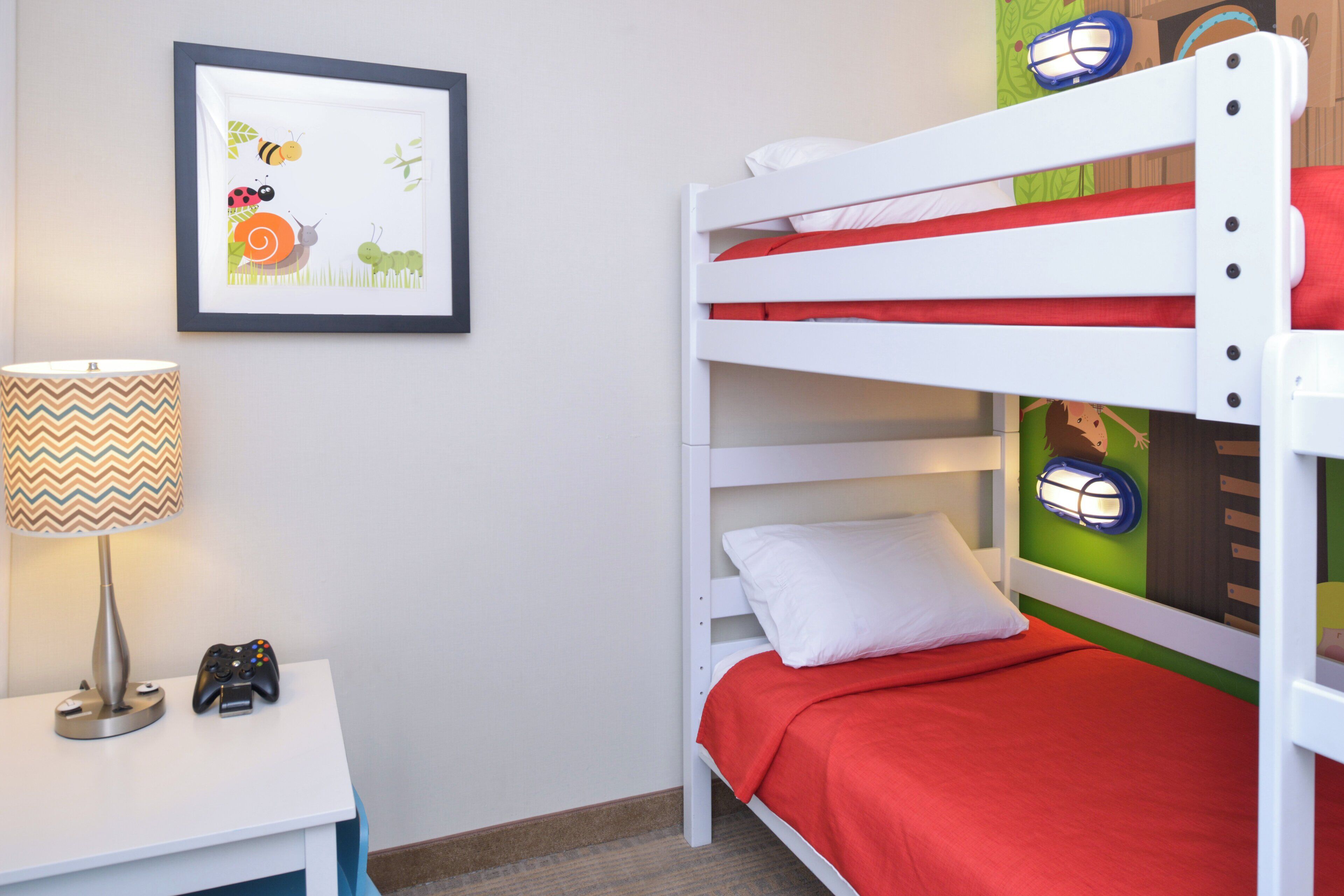 A bunk bed with a red bedspread, a framed picture on the wall, a lamp, and a gaming controller on a side table.
