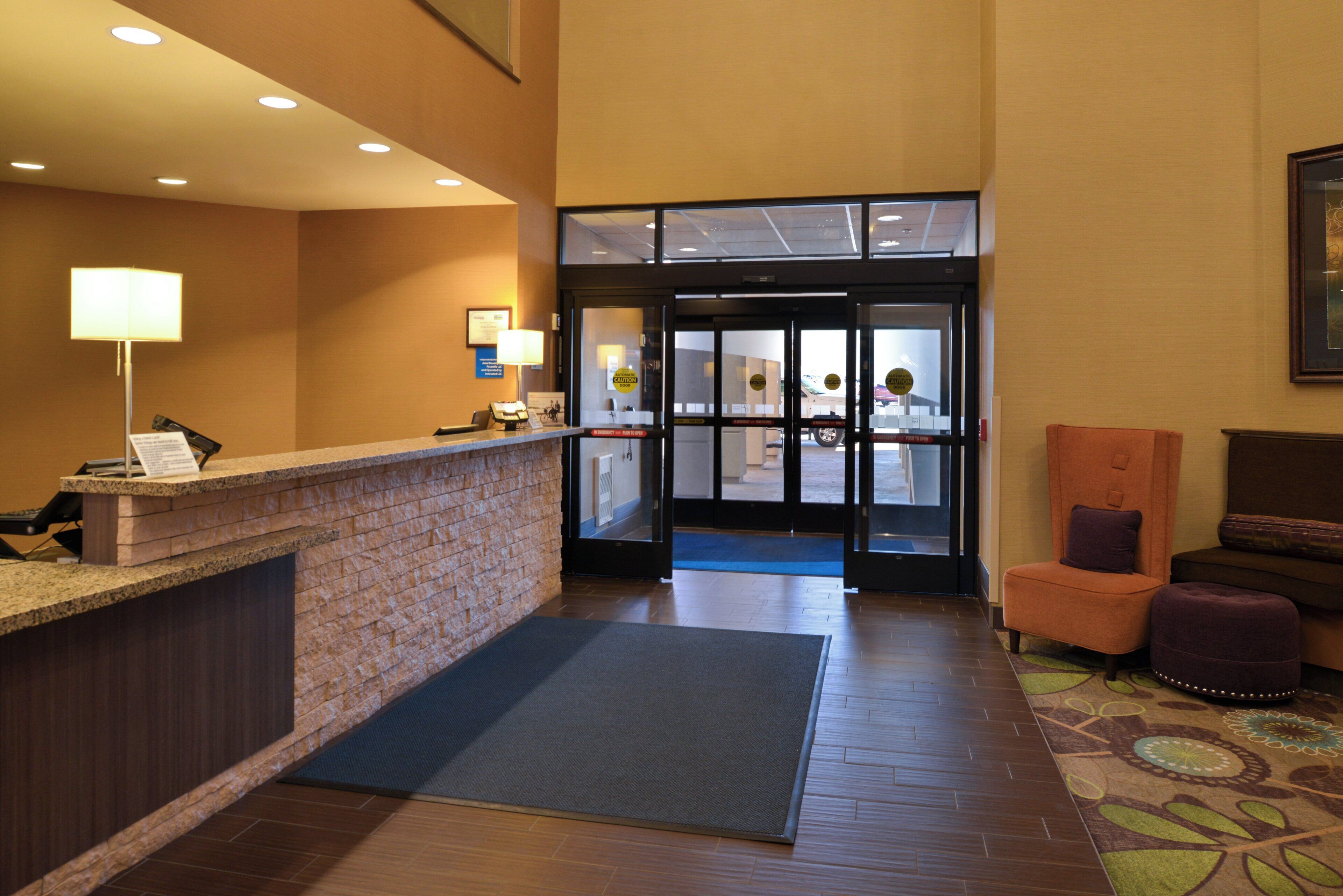 A hotel reception area with a reception desk, a seating area with a chair and ottoman, and a glass door entrance.
