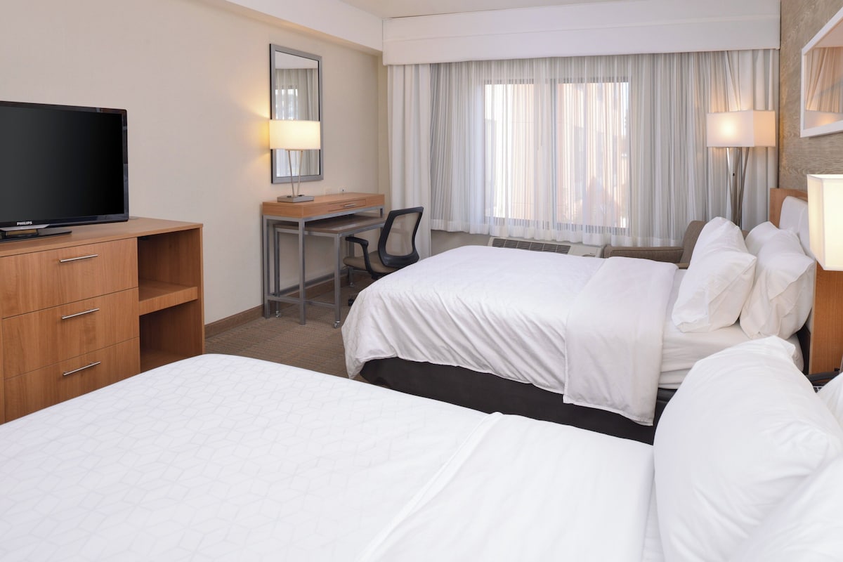 Une chambre d’hôtel avec deux lits, un bureau, une chaise, une télévision et une grande fenêtre avec des rideaux.